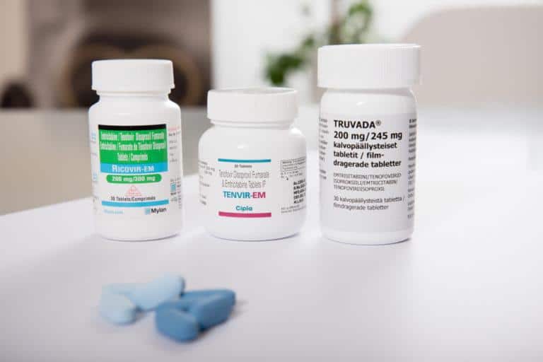 In the picture there are PReP medicines from different manufacturers. In the back there are three medicine bottles and in the front there are some blue pills. 