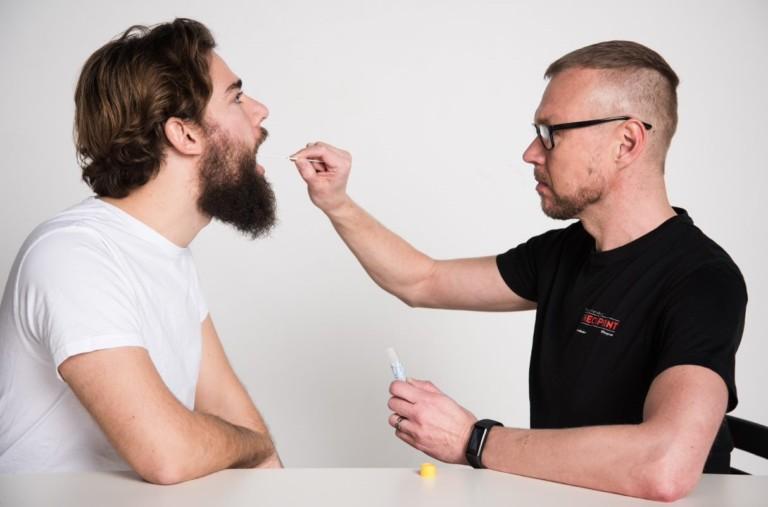 Two persons sitting next to table. One takes a sample from the other ones mouth.