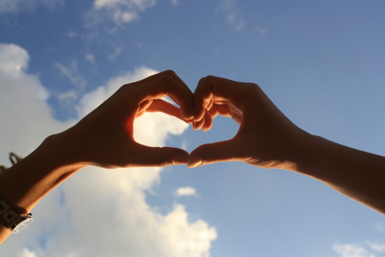 In the picture: two hands that form a shape of heart. In the background there is blue sky and clouds.