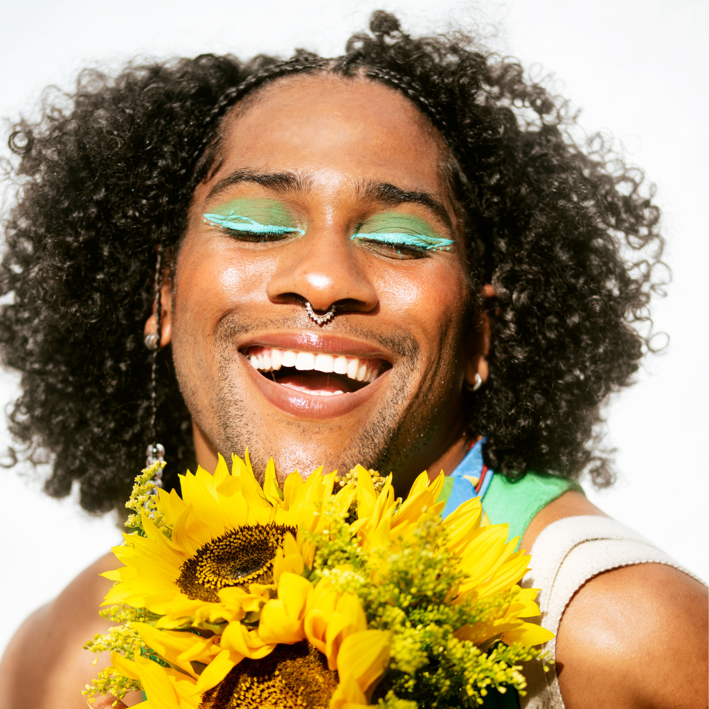 A smiling queer-presenting person with curly hair, bright green eye makeup, and a septum piercing, holding a bouquet of sunflowers against a white background.