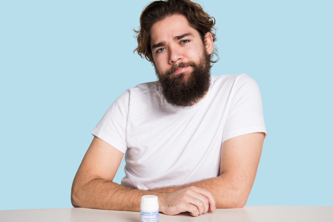 A white, masculine person is sitting at the table, looking reflective. On the table, there's a PrEP pill jar.