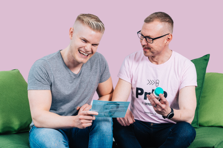 Two people are sitting on a sofa, smiling. One is holding a PrEP leaflet, the other one is holding a PrEP pill jar.